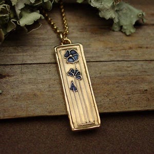 May include: Gold-tone rectangular pendant necklace featuring a floral design with blue enamel accents. The pendant hangs from a delicate gold chain. The necklace is set against a rustic wooden background.