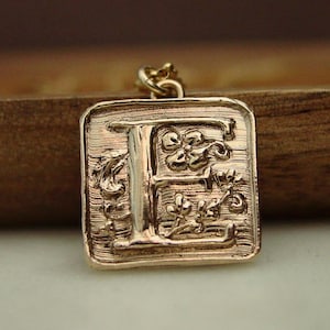 May include: A square gold-toned pendant featuring a raised letter E in the center. The surface of the pendant has a textured, brushed finish with embossed floral and vine details surrounding the letter. A small metal jump ring is attached to the top.