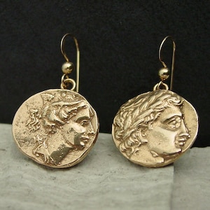May include: Gold-toned coin earrings featuring detailed profiles of classical figures. The earrings have a hook closure and a small ball accent. The coin pendants are round and have an aged appearance, suggesting an antique or vintage style.