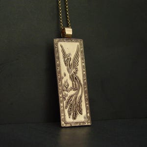 May include: A rectangular gold-toned pendant necklace featuring an embossed phoenix design with intricate wing and tail feather details. The pendant is framed by a decorative border and hangs from a matching gold-toned metal chain link necklace.