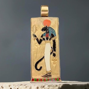 Puede incluir: Un colgante rectangular de tono dorado con una ilustración esmaltada de una deidad egipcia con cabeza de gato, que viste una falda blanca y un tocado azul, rematado con un disco solar rojo, sosteniendo un sistro y un ankh, con base roja y verde.