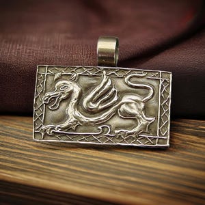 An antiqued rectangle bronze pendant with a wide loop on top for a chain to go through. On the pendant is an embossed Medieval style dragon crouching.
