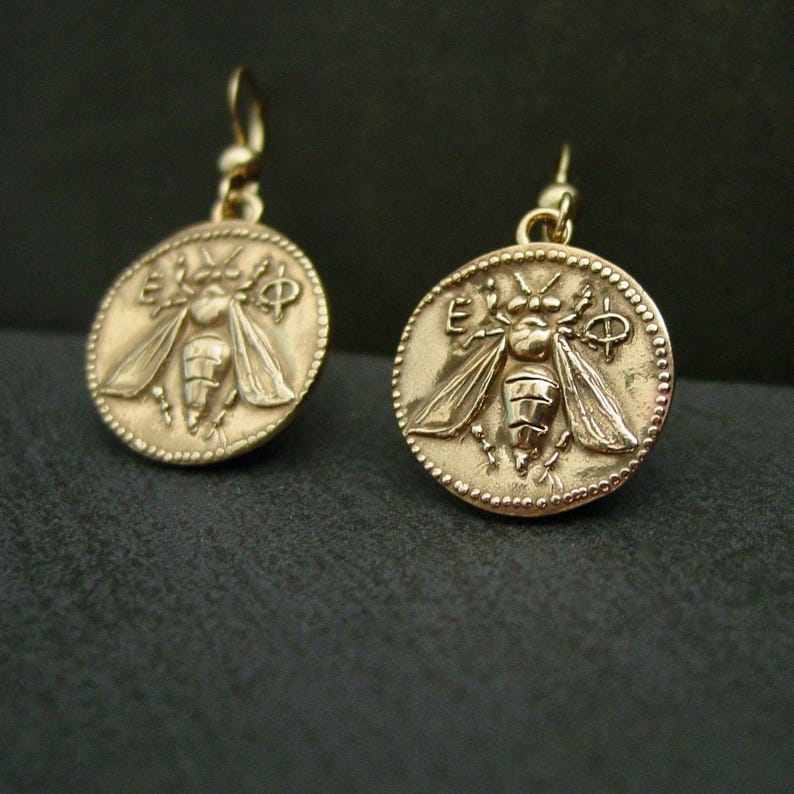 A pair of round gold coin earrings with dotted edge and a raised honey bee in its center.