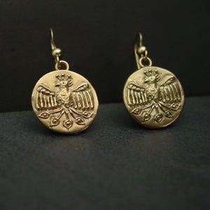 May include: A pair of gold-colored earrings. Each earring features a circular pendant with a detailed relief of a bird with outstretched wings. The bird is surrounded by decorative elements. The earrings have a simple hook closure.