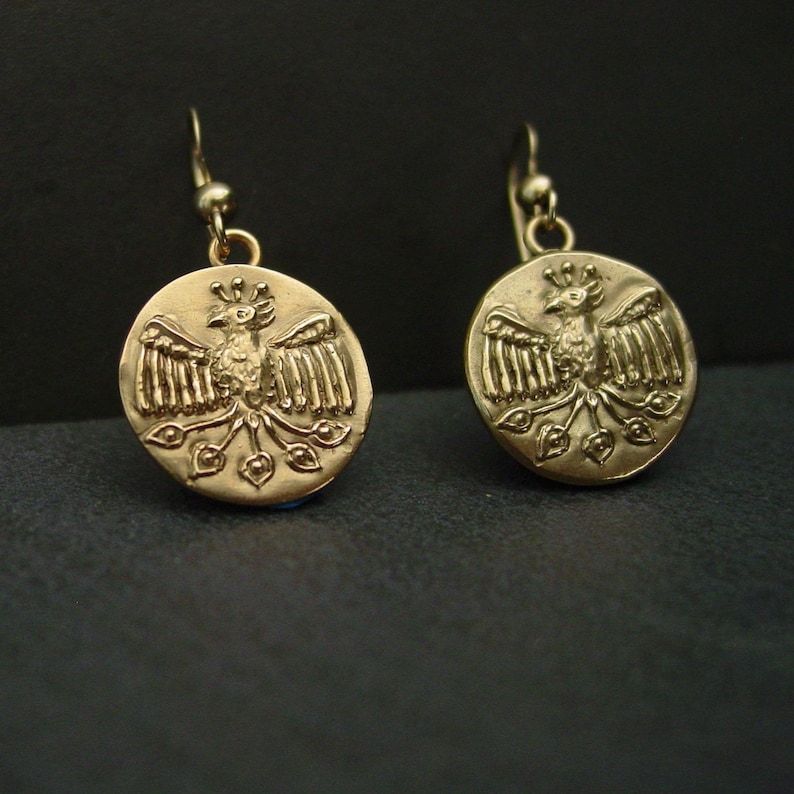 May include: A pair of gold-colored earrings. Each earring features a circular pendant with a detailed relief of a bird with outstretched wings. The bird is surrounded by decorative elements. The earrings have a simple hook closure.