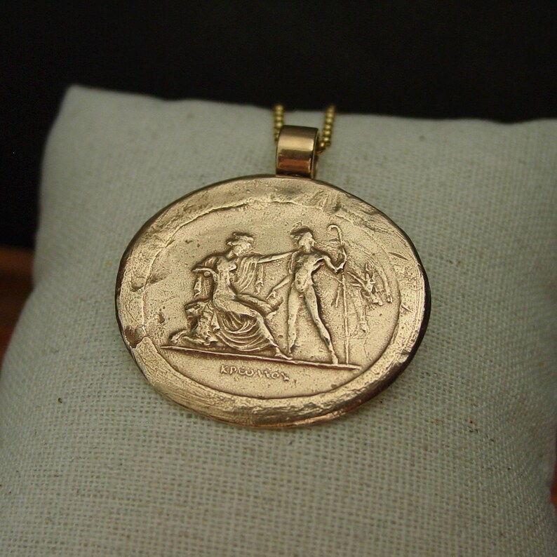 May include: A gold-colored pendant with a detailed relief of classical figures. The round pendant hangs from a delicate gold chain. The relief depicts two figures and text at the bottom. The pendant is set against a white background.