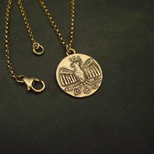 May include: A gold-toned necklace featuring a circular pendant with a raised relief of a stylized bird with spread wings and a fanned tail. The pendant hangs from a delicate gold-toned rolo chain that includes a lobster claw clasp closure.
