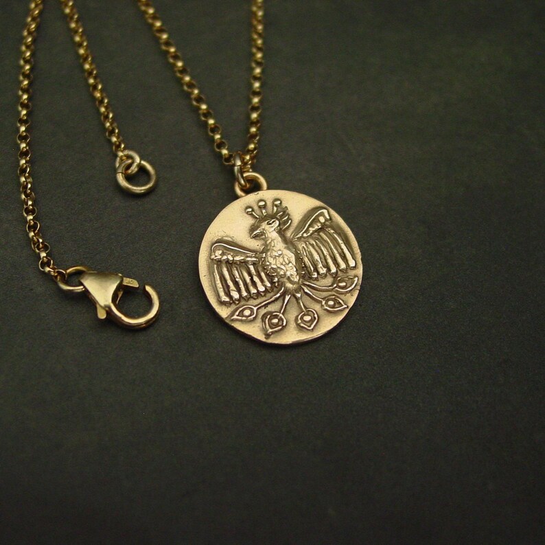 May include: A gold-toned necklace featuring a circular pendant with a raised relief of a stylized bird with spread wings and a fanned tail. The pendant hangs from a delicate gold-toned rolo chain that includes a lobster claw clasp closure.