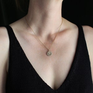 May include: A gold-toned necklace featuring a delicate chain and a small, circular pendant. The pendant displays an embossed eagle design with spread wings, centered on the round metal disc. The necklace rests against the neck, showcasing the metal finish.