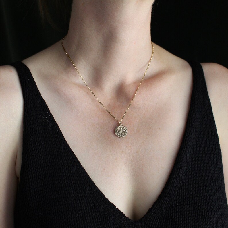 May include: A gold-toned necklace featuring a delicate chain and a small, circular pendant. The pendant displays an embossed eagle design with spread wings, centered on the round metal disc. The necklace rests against the neck, showcasing the metal finish.