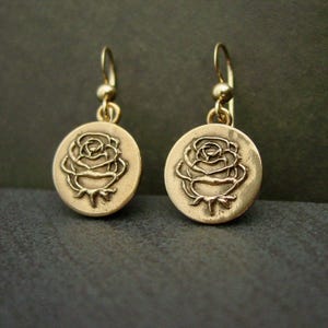 May include: A pair of gold-colored earrings. Each earring features a circular pendant with a raised rose design. The earrings have a simple hook closure. The background is a dark, neutral color.