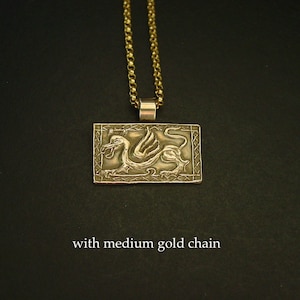 K&ouml;nnte beinhalten: Goldene Halskette mit Anh&auml;nger, der ein detailliertes gefl&uuml;geltes Drachenmotiv auf einer rechteckigen Plakette zeigt. Die Halskette ist mit einer mittelgro&szlig;en goldenen Kette kombiniert.