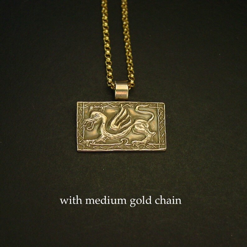 K&ouml;nnte beinhalten: Goldene Halskette mit Anh&auml;nger, der ein detailliertes gefl&uuml;geltes Drachenmotiv auf einer rechteckigen Plakette zeigt. Die Halskette ist mit einer mittelgro&szlig;en goldenen Kette kombiniert.