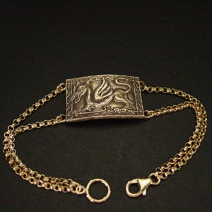 May include: A gold bracelet with a rectangular pendant featuring a detailed embossed design of a winged dragon. The bracelet has a double chain design with a lobster clasp and a circular ring.