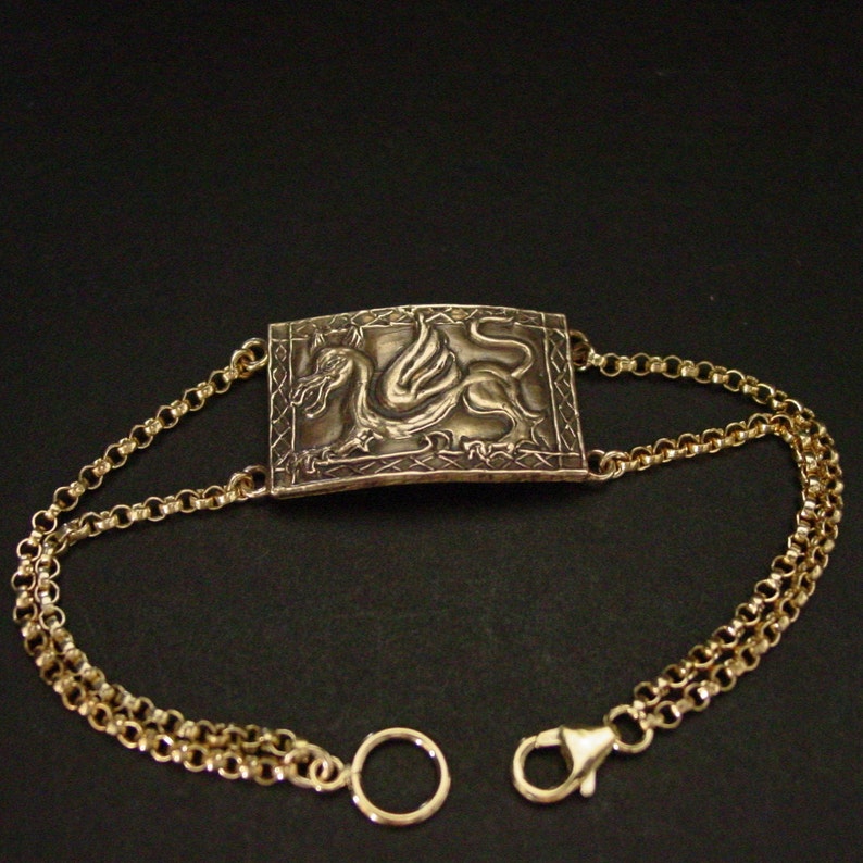 May include: A gold bracelet with a rectangular pendant featuring a detailed embossed design of a winged dragon. The bracelet has a double chain design with a lobster clasp and a circular ring.