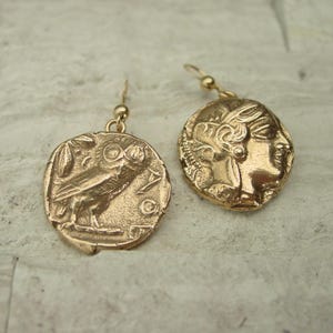 May include: Gold-colored coin earrings. One earring features an owl, the other a profile of a woman. The earrings have a hook closure. The coins have an antique, textured appearance.