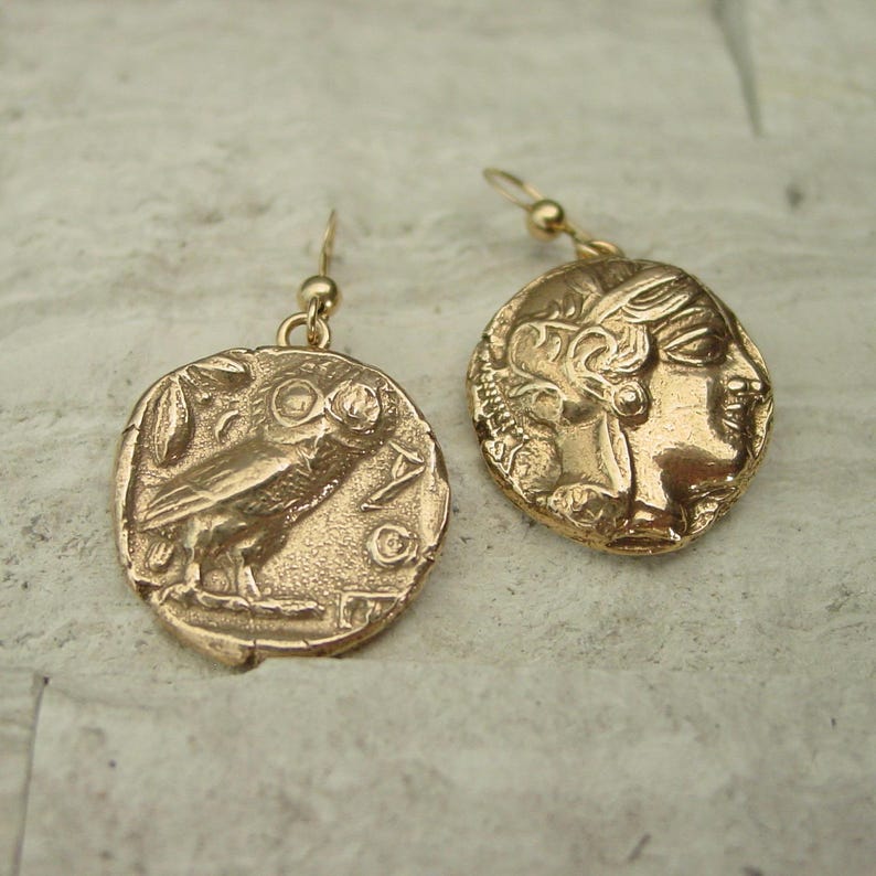 May include: Gold-colored coin earrings. One earring features an owl, the other a profile of a woman. The earrings have a hook closure. The coins have an antique, textured appearance.