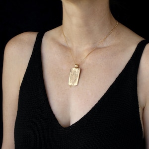 May include: A gold-toned rectangular pendant necklace featuring an intricate engraved design on the front surface. The pendant hangs from a delicate gold-toned chain, resting against a black textured fabric garment with a deep V-neckline.