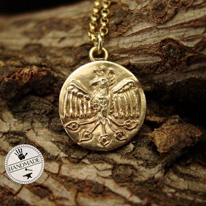 A gold round coin hanging on a thin gold rolo style chain. On the pendant is a raised Phoenix with spread wings.