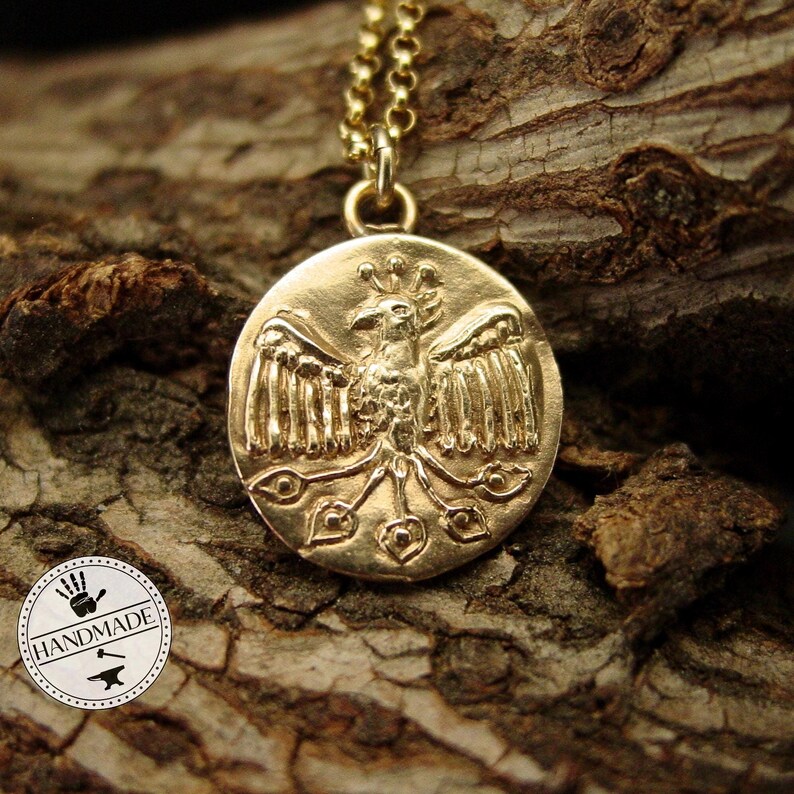 A gold round coin hanging on a thin gold rolo style chain. On the pendant is a raised Phoenix with spread wings.