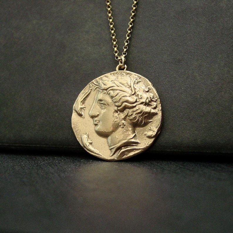 A large gold round coin hanging from a thin gold link chain. On the pendant is a raised profile of a woman with her wavy hair up in a hairdo. She is wearing some drop crotalia earrings and a pearl necklace. She is facing two frolicking dolphins.