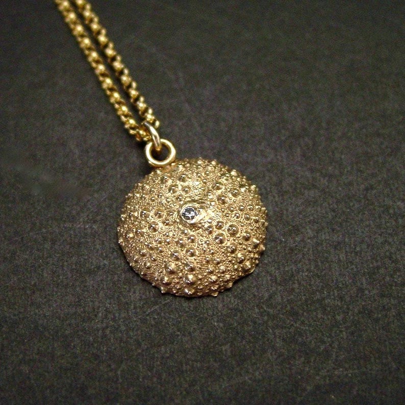 A gold round sea urchin shell charm with lots of texture hanging from a thin gold Rolo style chain. In its center is a white sparkly clear stone. The necklace is dainty.