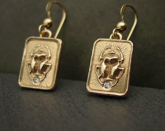 May include: A pair of gold-colored earrings. Each earring features a rectangular pendant with a detailed scarab beetle design and a small, clear gemstone. The earrings have a hook closure.