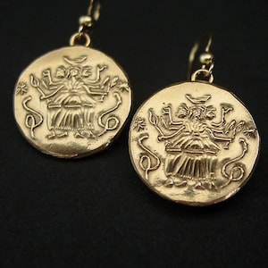 May include: A pair of gold-toned drop earrings featuring circular coin-shaped pendants. Each pendant displays an embossed design of a multi-armed figure seated on a throne, flanked by two snake-like forms and small star symbols, suspended from hooks.