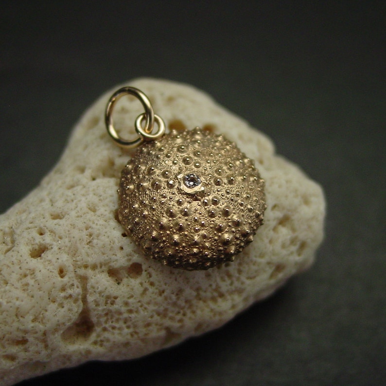 May include: A gold-coloured pendant shaped like a sea urchin, with a small gemstone set in the centre. The pendant hangs from a gold loop. The pendant is displayed on a piece of light-coloured coral.