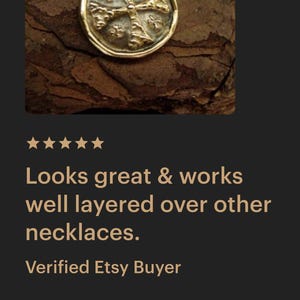 May include: A gold-toned circular pendant necklace featuring a raised cross design with textured details. The pendant is suspended from a delicate gold-toned chain, showcasing a metallic finish and a coin-like shape with a slightly irregular, rounded edge.