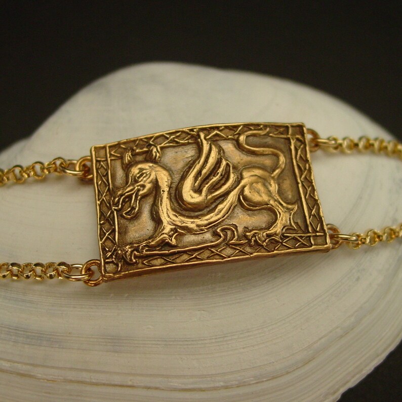 May include: A gold chain bracelet with a rectangular pendant featuring a detailed embossed image of a winged griffin.