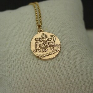 May include: A gold-toned circular pendant necklace featuring an embossed mythological figure riding a creature. The pendant hangs from a delicate gold-toned ball chain, showcasing a textured surface with a decorative border along the bottom edge.