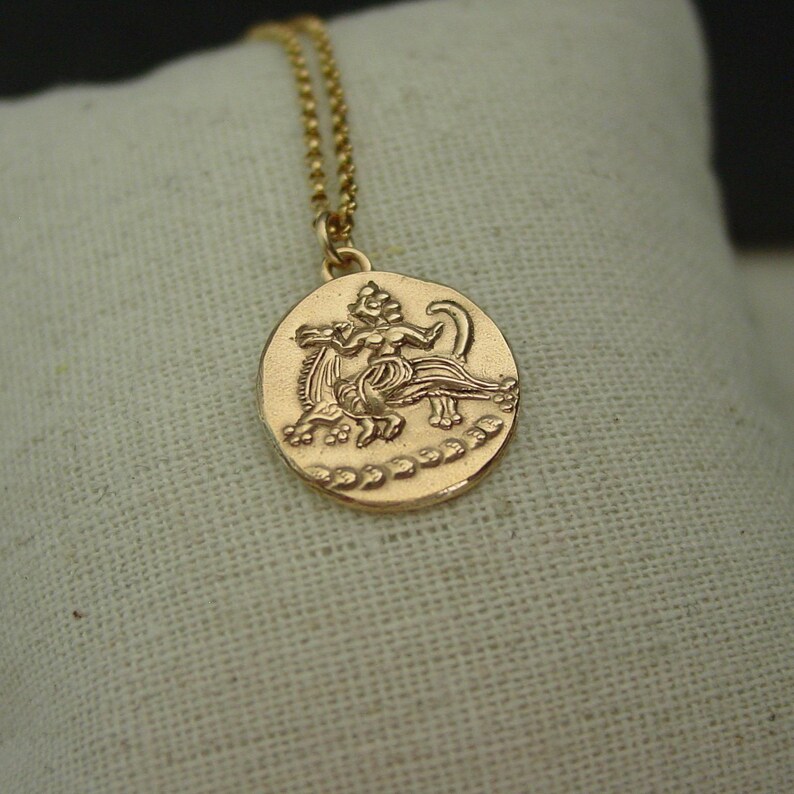 May include: A gold-toned circular pendant necklace featuring an embossed mythological figure riding a creature. The pendant hangs from a delicate gold-toned ball chain, showcasing a textured surface with a decorative border along the bottom edge.