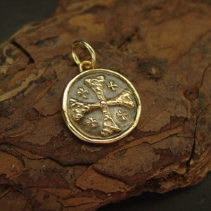 May include: A circular gold pendant featuring a textured cross design centred on a dark, recessed background. The pendant has a polished gold rim and a small gold jump ring attached to the top for hanging, showcasing a detailed, coin-like appearance.