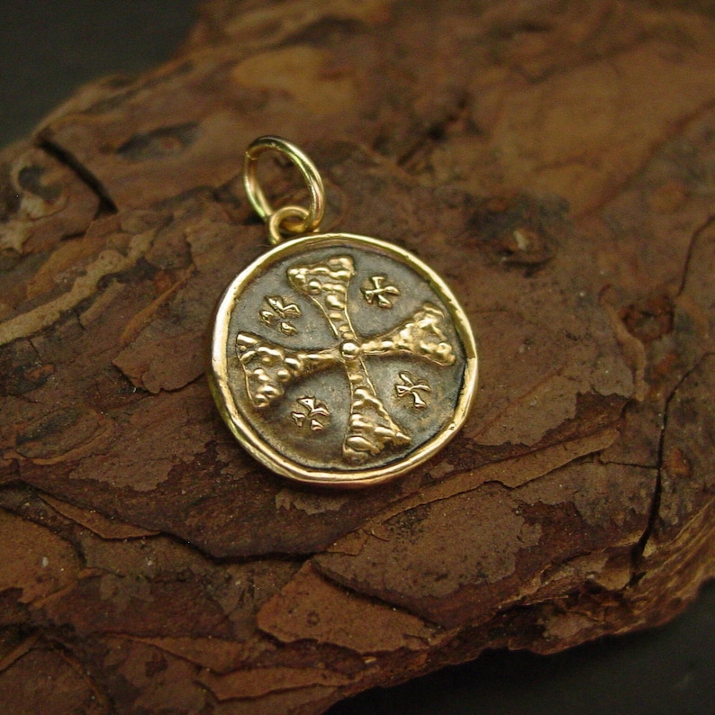 May include: A circular gold pendant featuring a textured cross design centred on a dark, recessed background. The pendant has a polished gold rim and a small gold jump ring attached to the top for hanging, showcasing a detailed, coin-like appearance.