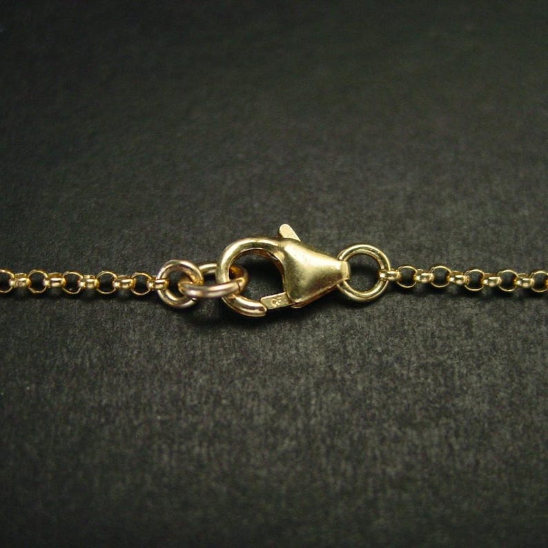 May include: Close-up of a gold-coloured necklace clasp and chain. The lobster clasp is triangular and attached to a delicate chain with small, round links. The background is a dark, solid colour.