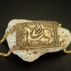 A gold rectangle horizontal bracelet. The center panel has an embossed dragon with a Medieval design frame around it. On each corners of the charm is attached a chain with then clasps in the back. The bronze panel is curved to follow the shape of the wrist.