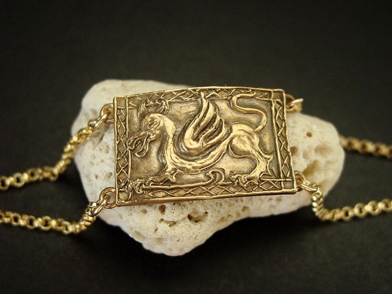 A gold rectangle horizontal bracelet. The center panel has an embossed dragon with a Medieval design frame around it. On each corners of the charm is attached a chain with then clasps in the back. The bronze panel is curved to follow the shape of the wrist.