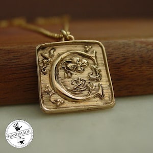 以下が含まれることがあります： 金色のペンダントネックレス。長方形のペンダントには、装飾的な文字「C」のデザインが施されています。ペンダントはテクスチャード加工の背景と花のアクセントがあります。ネックレスは手作りです。