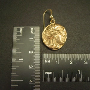 May include: A gold-colored earring with a circular pendant featuring a profile of a person. The earring has a hook closure. The pendant is approximately 1 inch in diameter.
