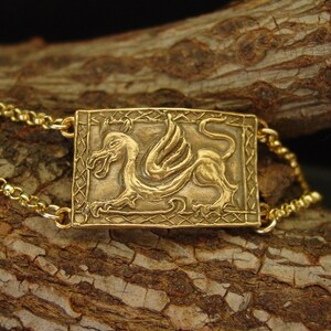 May include: A gold-toned bracelet with a rectangular pendant featuring a detailed embossed winged dragon design.