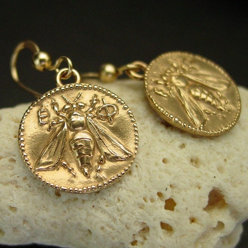 Small round gold earrings with a bee in its center and Greek letters with a dotted frame.
