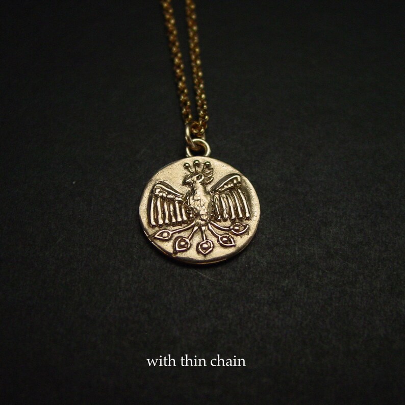 May include: A gold-toned circular pendant necklace featuring a relief design of a bird with spread wings and a fanned tail. The pendant is suspended from a delicate, thin gold-toned chain. The text "with thin chain" is printed at the bottom.