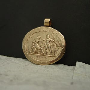 May include: A gold-colored pendant with a detailed relief of classical figures. The round pendant features a scene with two figures and a staff, with a loop for a chain at the top. The background is dark.