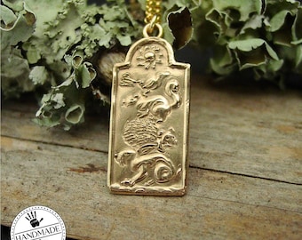 May include: A gold-colored pendant necklace with a rectangular pendant featuring a detailed relief of a mythical creature, birds, and sun. The necklace is on a gold chain. The background includes a handmade logo and natural elements.