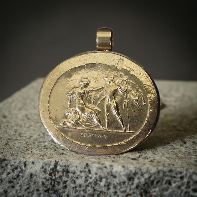 A gold horizontal oval pendant with a wide loop on top for a chain. On the pendant is a scene in relief of the god Hermes standing in front of Persephone seated on her throne next to a lion.
