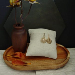 May include: Gold-colored coin earrings with an eagle design are displayed on a cream-colored pillow. The earrings are on a wooden tray with a small brown vase and dried flower petals. The background is dark.