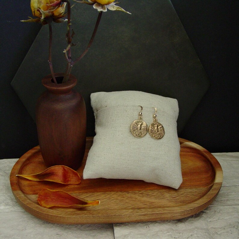May include: Gold-colored coin earrings with an eagle design are displayed on a cream-colored pillow. The earrings are on a wooden tray with a small brown vase and dried flower petals. The background is dark.