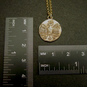 May include: A gold-toned circular pendant featuring a relief of a bird with spread wings and a fanned tail, suspended from a matching chain. The pendant is positioned next to two black rulers, showing a diameter of approximately 0.75 inches.