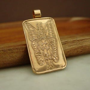 May include: A rectangular gold-toned pendant featuring an engraved illustration of a winged figure with antennae. The pendant has a smooth, raised border and a small bail at the top for attachment. The design depicts a humanoid form with large, detailed butterfly-like wings extending from the back, rendered in a metallic finish.
